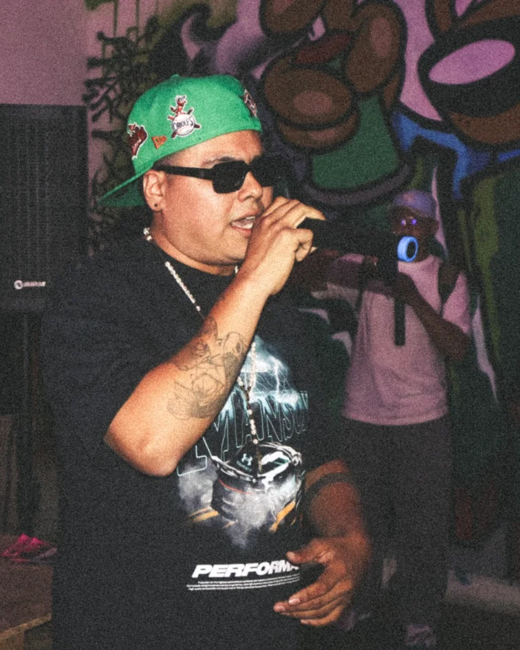 El Gordo Wichi cantando en vivo, con gorra verde y lentes oscuros, frente a un mural